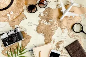 A flat lay composition of travel-related items on top of a world map, including an open notebook with a pen, a vintage camera, a straw hat, sunglasses, a smartphone, and earphones, suggesting planning or journaling for a blogging journey.