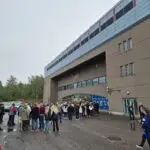 Group of individuals walking towards an icehockey areena with a blue and white facade.