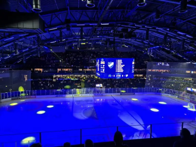Indoor ice hockey arena with a rink illuminated in blue light, filled stands, and a large scoreboard above the center, indicating an exciting game atmosphere between kiekko-espoo - hifk.