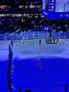 Indoor ice hockey rink with a blue hue, several people standing near the center while giving a speech, and a the hometeam players gathered by the edge of the rink in a straight line