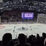 Indoor ice hockey arena during a game, viewed from the stands, with the rink and players in the middle distance, well-lit arena, and many spectators in the stands. A large scoreboard is visible above the rink.