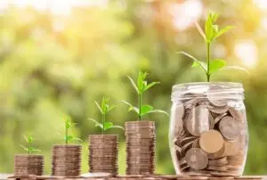 A series of coins stacked in ascending order with small plants growing out of the top of each stack, symbolizing financial growth and investment over time. The largest stack is inside a glass jar, representing savings and wealth accumulation.
