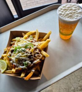 Tray with loaded fries topped with cheese, green onions, sauce, and meat, served with a fizzy drink.