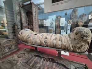 "Mummy of Cleopatra from the Roman Period, displayed in a museum, wrapped in linen bandages and placed inside a decorated sarcophagus, surrounded by artifacts and informational displays.