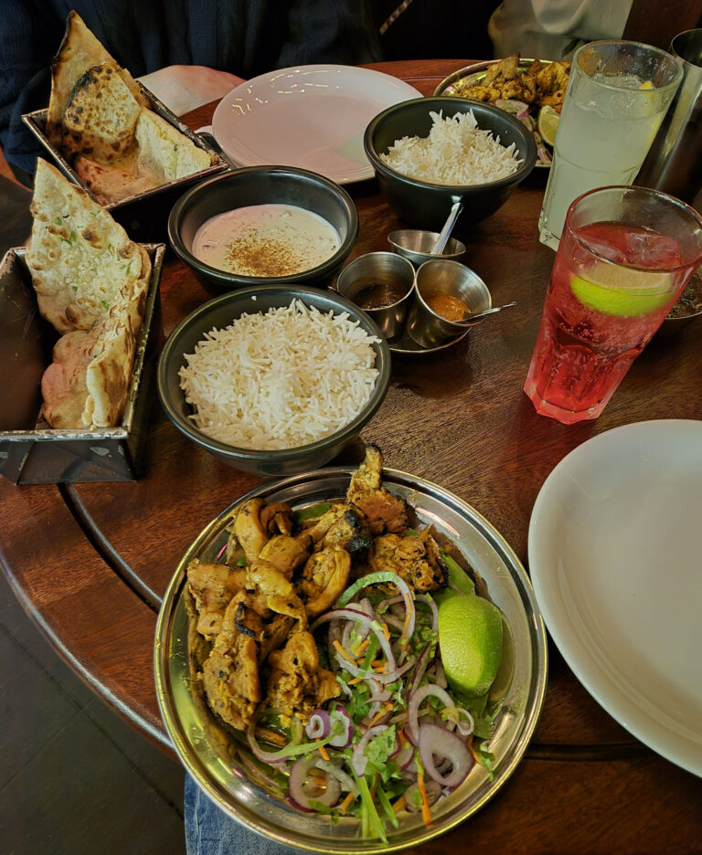 Table set with various Indian dishes from Dishoom, including naan bread, rice, raita, grilled chicken, condiments, and beverages.