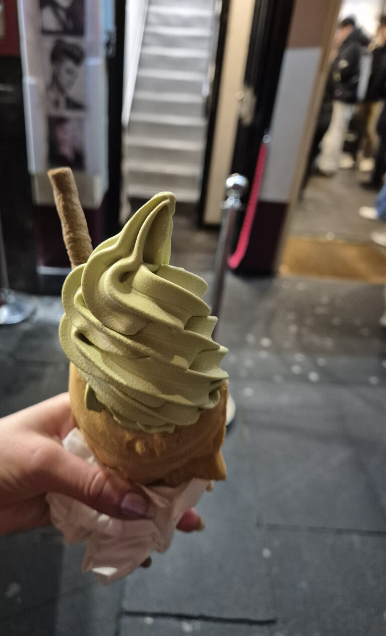 A hand holding a matcha-flavored soft-serve ice cream in a fish-shaped taiyaki cone, with a wafer stick on top, in Chinatown, London.