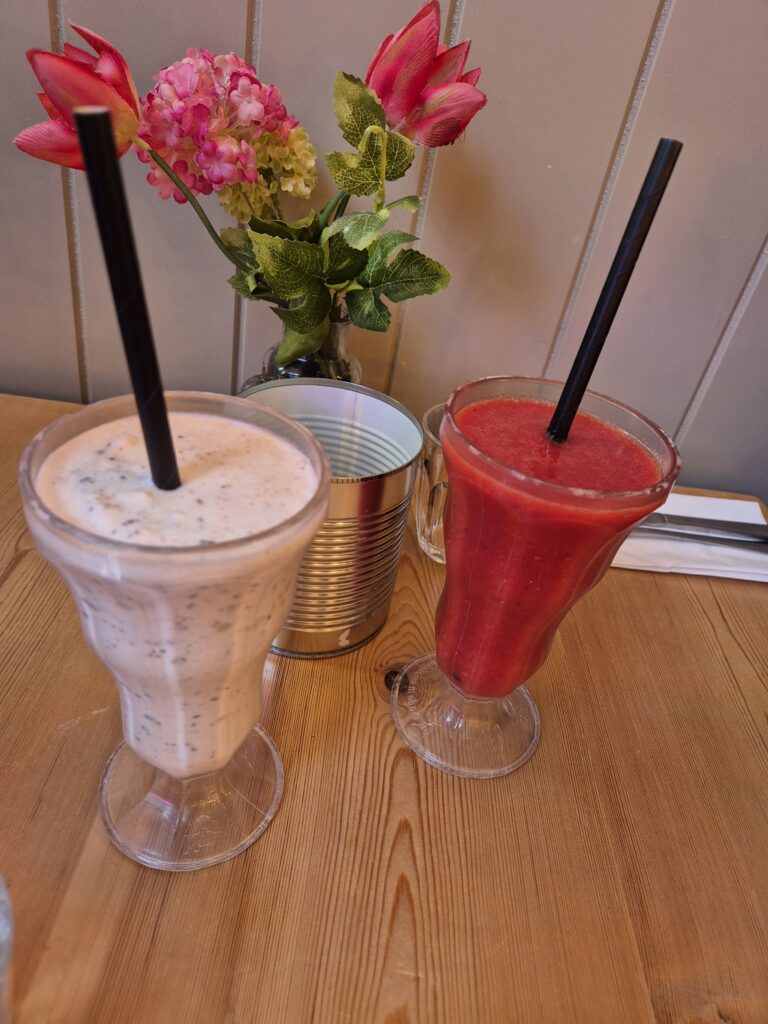 Two beverages on a wooden table: a red berry smoothie and a Kinder Bueno milkshake, served in tall glasses with black straws, with artificial flowers in a tin can.