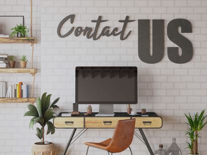 A modern home office setup with a computer on a desk, a chair, and decorative plants. The words "Contact US" are prominently displayed on the wall above the desk.
