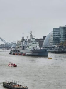 HMS Belfast, a large naval ship, docked at a pier, and a small red boat and buildings in the foreground.