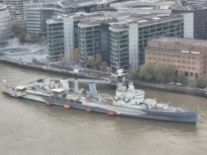 Aerial view of HMS Belfast, a large grey naval warship, docked on a river with modern buildings in the background.