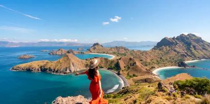 A person in a red dress and wide-brimmed hat stands on a hill overlooking a scenic coastal landscape with multiple bays and rugged terrain.