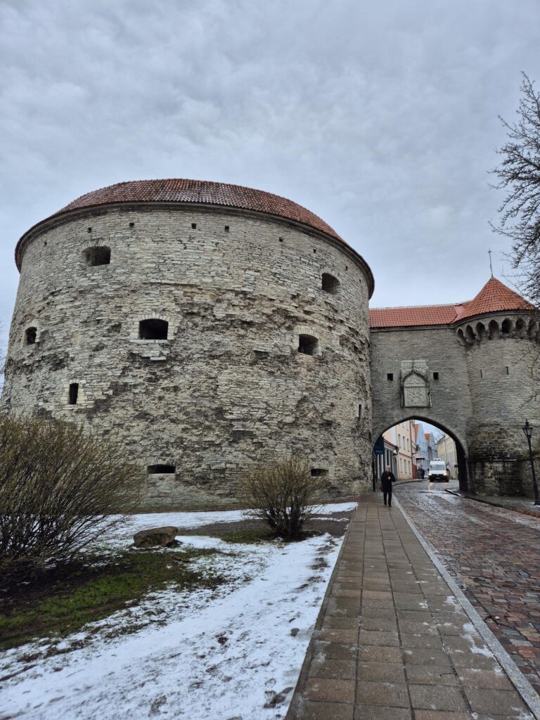 Medieval stone tower in Tallinn Old Town with a red-tiled roof and an archway, surrounded by snow and greenery.