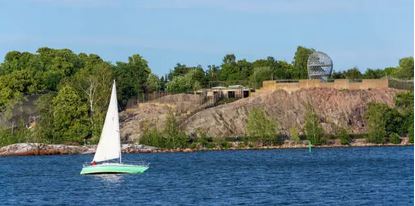 A sailboat glides across the water near Korkeasaari Helsinki Zoo, with a tree-covered shoreline and rocky outcrop in the background.