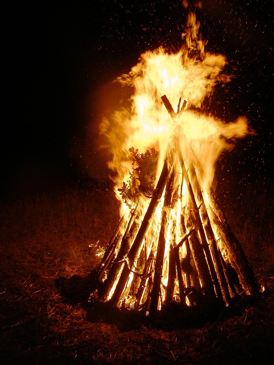 Towering midsummer bonfire blazing against the night sky, with orange flames lighting up the darkness and casting a warm glow on the surroundings.