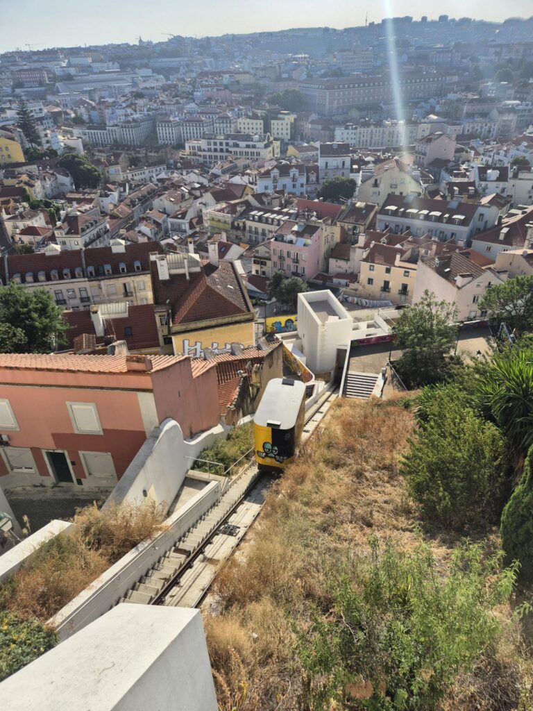 Yellow and white funicular ascending a track lined with greenery and buildings, heading toward a miradouro (viewpoint) in Lisbon.