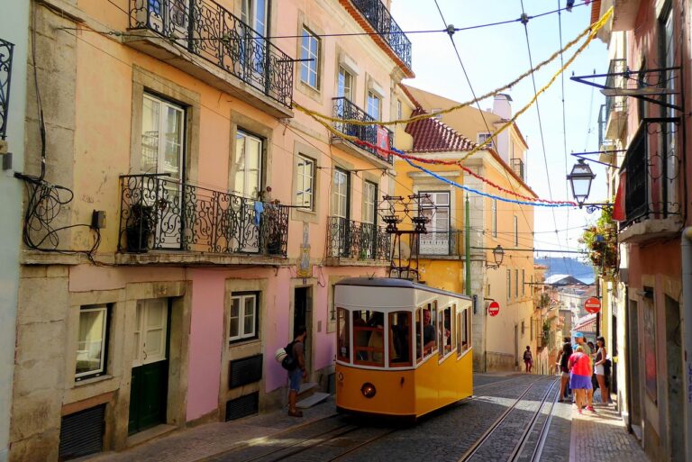 Lisbon’s iconic yellow tram/ funicular traveling up a cobblestone street lined with pastel buildings and colorful streamers.