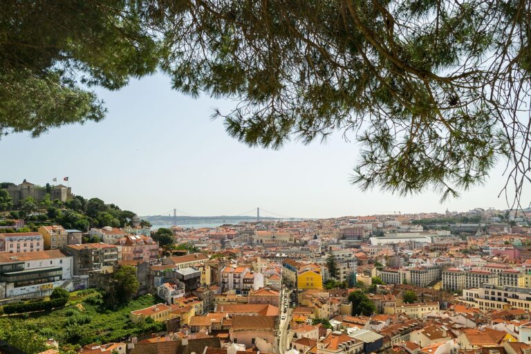 Scenic view from Miradouro da Graça in Lisbon, with red-tiled rooftops, the 25 de Abril Bridge in the background, and tree branches framing the top.