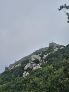A distant tower of the Moorish Castle in Sintra, surrounded by mist, trees, and rocky terrain.