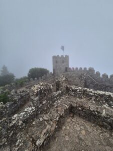 A stone tower of the Moorish Castle in Sintra, partially veiled in mist atop a forested hill.
