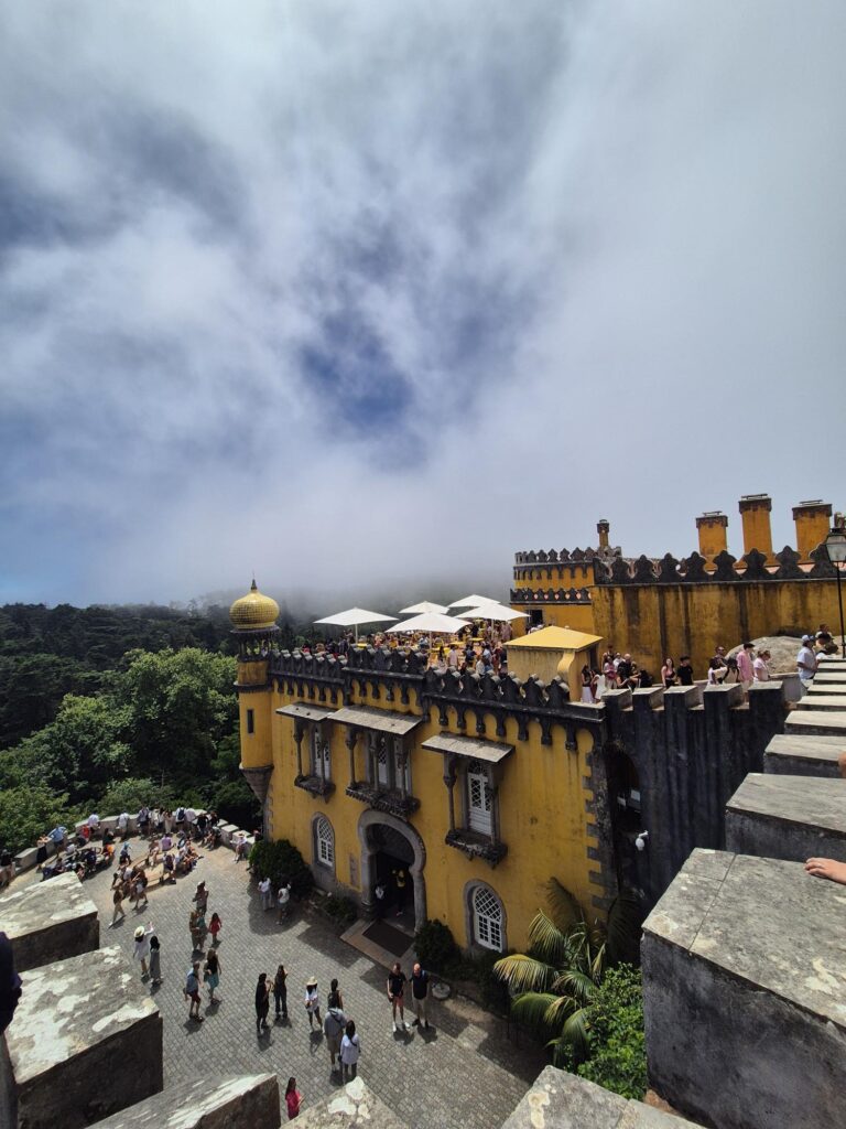A view of Pena Palace in Sintra, showing its vibrant yellow walls, multiple turrets, and a terrace filled with visitors. The background includes lush greenery and a cloudy sky. One of Sintra castles.