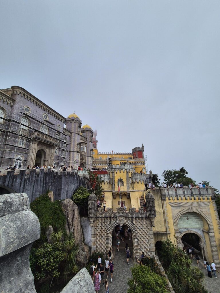 Wide view of Pena Palace with yellow and red sections, surrounded by greenery and perched on a hill.