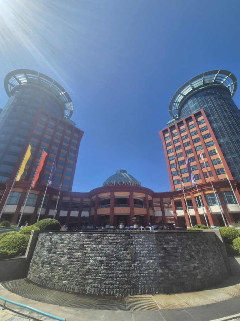 Colombo Mall, the best shopping malls in Lisbon Portugal. Featuring Colombo Mall, a modern building with two cylindrical towers on either side and a central section with flags hanging from it. The sky is clear and blue, and sunlight is shining brightly.