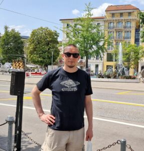Blog writer Mathias MysteryWriter at Lugano, Switzerland wearing a black t-shirt and beige shorts. There is a fountain and a large building with trees around it in the background.