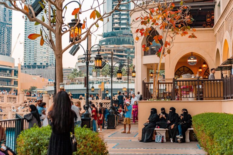 Outdoor terrace and promenade in a modern Middle Eastern city, featuring traditional-style architecture with arched doorways, ornate lanterns, and decorative railings, surrounded by high-rise buildings in the background and people walking or sitting along the walkway.