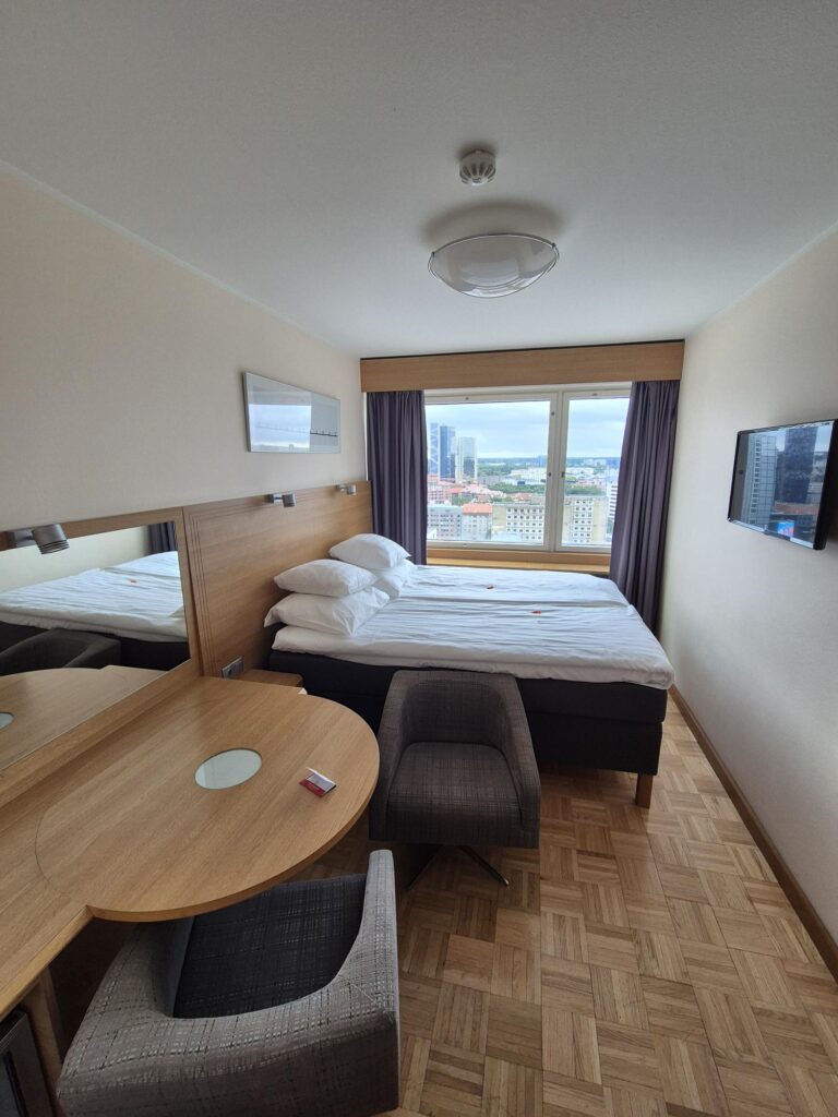 Hotel room on the 21st floor of Sokos Hotel Viru, featuring a double bed, a round wooden table with two chairs, and a large window offering a panoramic city view. The room has light-colored walls and parquet flooring. A Sokos Hotel Viru Tallinn Review.