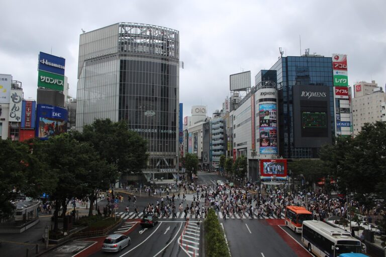 Busy urban intersection in a modern city with large glass buildings, colorful billboards, and crowds of pedestrians crossing multiple crosswalks under an overcast sky.