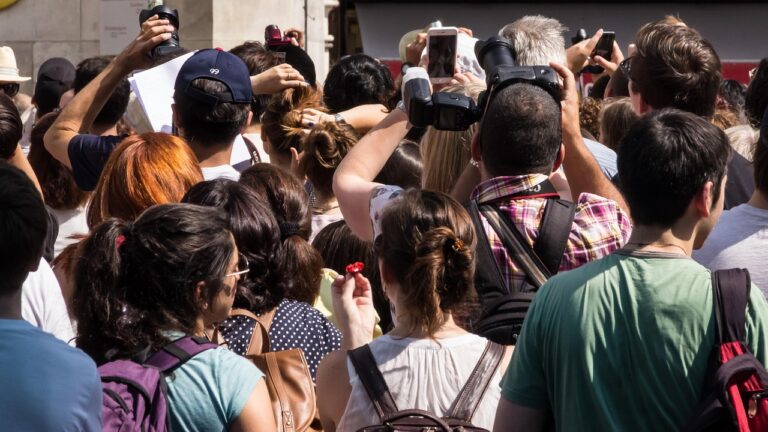 Crowd of people gathered outdoors, many holding cameras and smartphones above their heads to take photos, with individuals wearing backpacks and casual clothing under bright sunlight. Travel Behavior Forecasts by Continent.