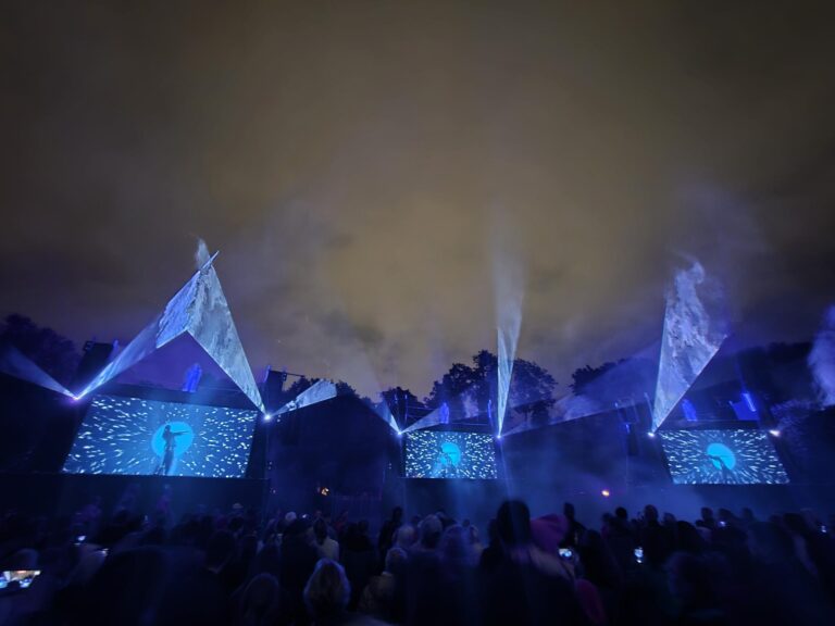 Outdoor stage at Valgus Kõnnib 2025 Light Festival illuminated with blue-toned projections and triangular light beams against a cloudy night sky, featuring large screens displaying abstract visuals and a silhouetted performer, with a crowd watching in the foreground.