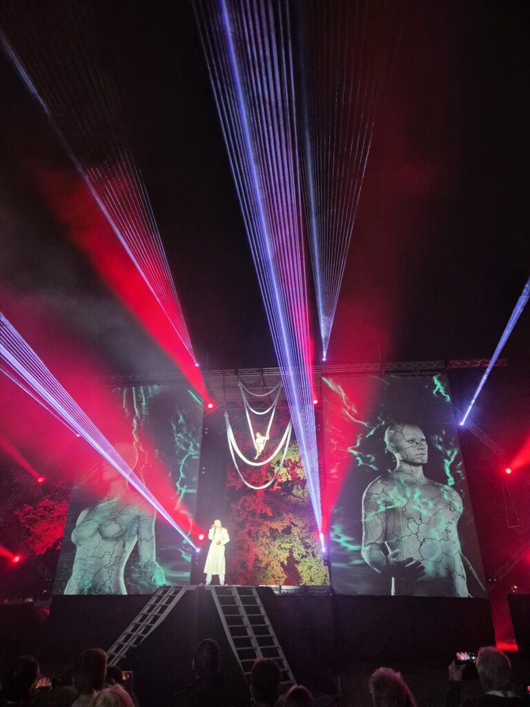 Stage performance at Valgus Kõnnib 2025 Light Festival featuring vivid red and blue laser beams cutting through the dark night sky, with large screens displaying abstract visuals of cracked stone-like figures and colorful patterns behind a performer in white attire.
