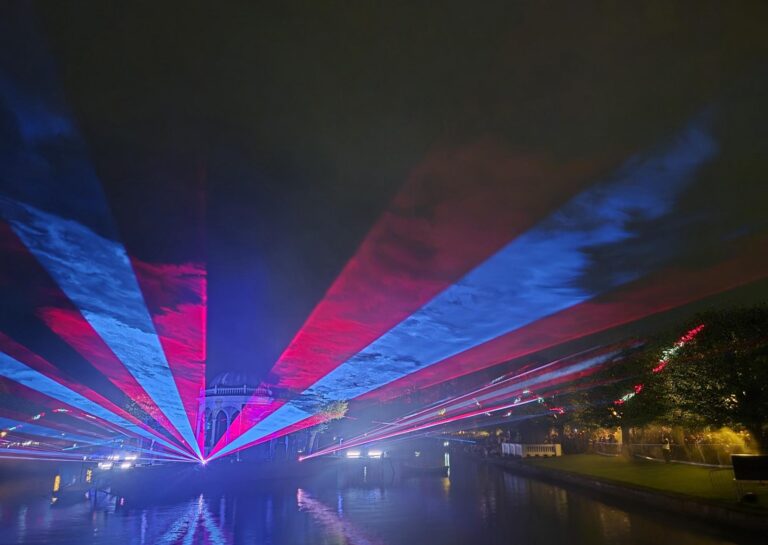 Colorful laser beams in red and blue fan out across a cloudy night sky above a water canal during Valgus Kõnnib 2025 Light Festival, with reflections shimmering on the water and a pavilion faintly visible in the background.