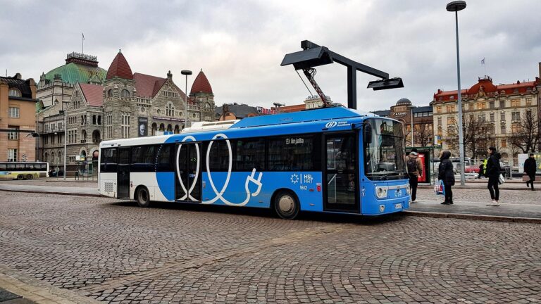 Blue electric bus charging at a station in Helsinki with historic buildings in the background.