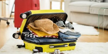 Yellow suitcase overflowing with clothes, a straw hat, shoes, and travel items, illustrating what happens when efficient packing tips for every type of trip are not followed.