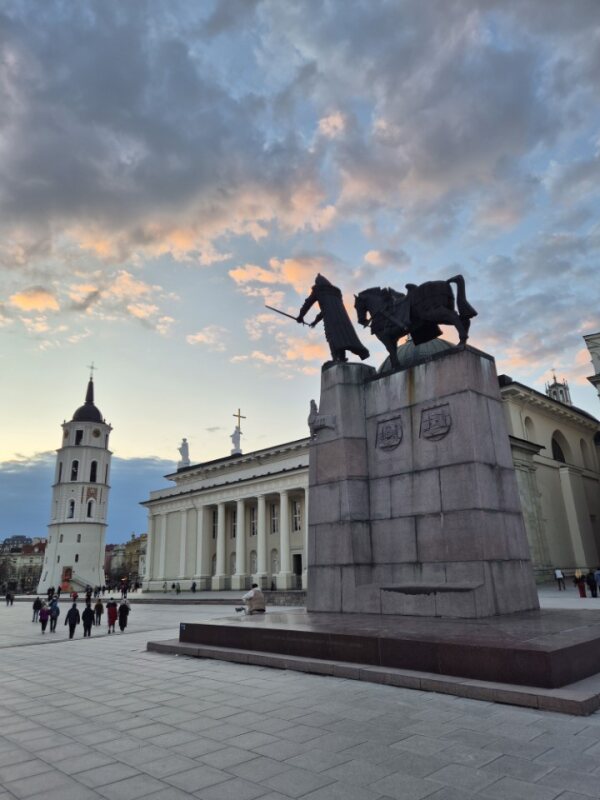 Large statue in a city square with a bell tower and cathedral